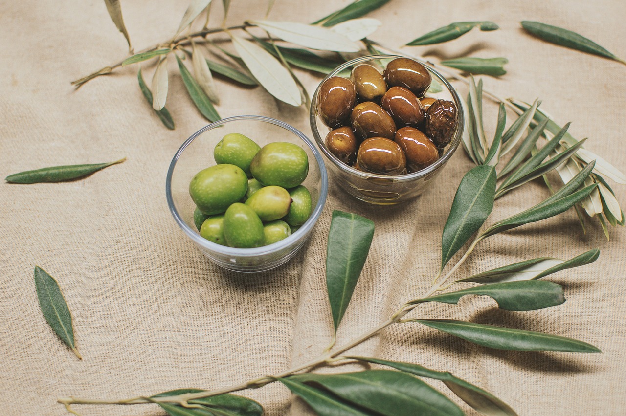 Fotografia de duas taças de azeitonas, uma de castanhas e outra de verdes, em cima de uma mesa decorada de ramos de oliveiras